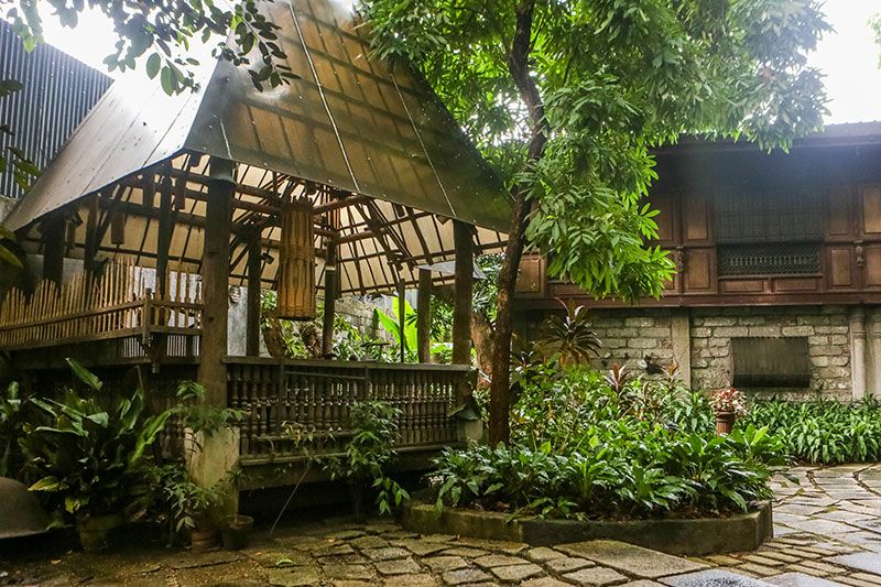 A bamboo and thatch bahay kubo built without nails standing in a rain-soaked garden, beside the capiz windows and stone wall of the main house