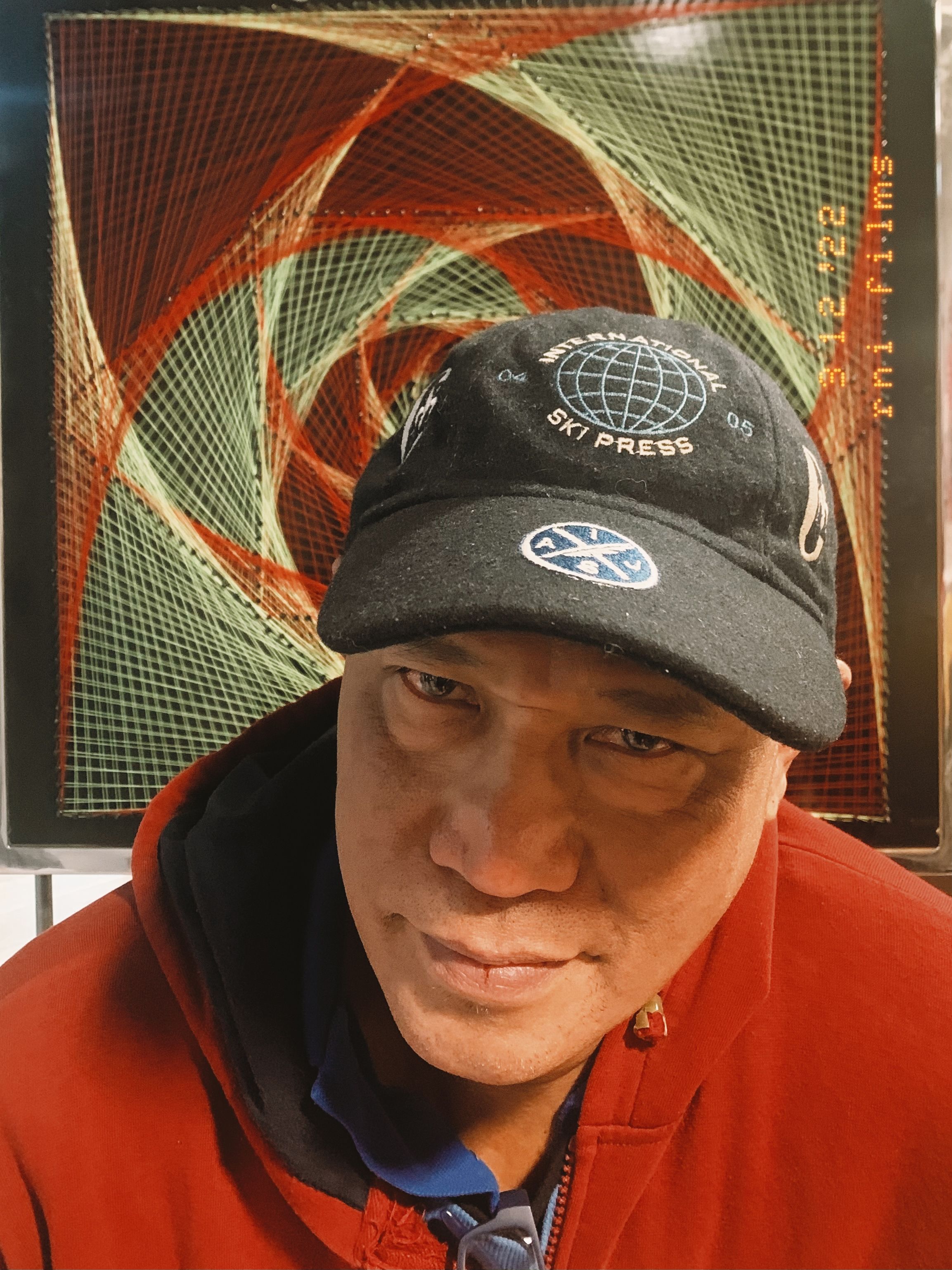 Esmundo Mamon in a red hoodie and an ‘International Ski Press’ cap before a red-and-green string-art panel