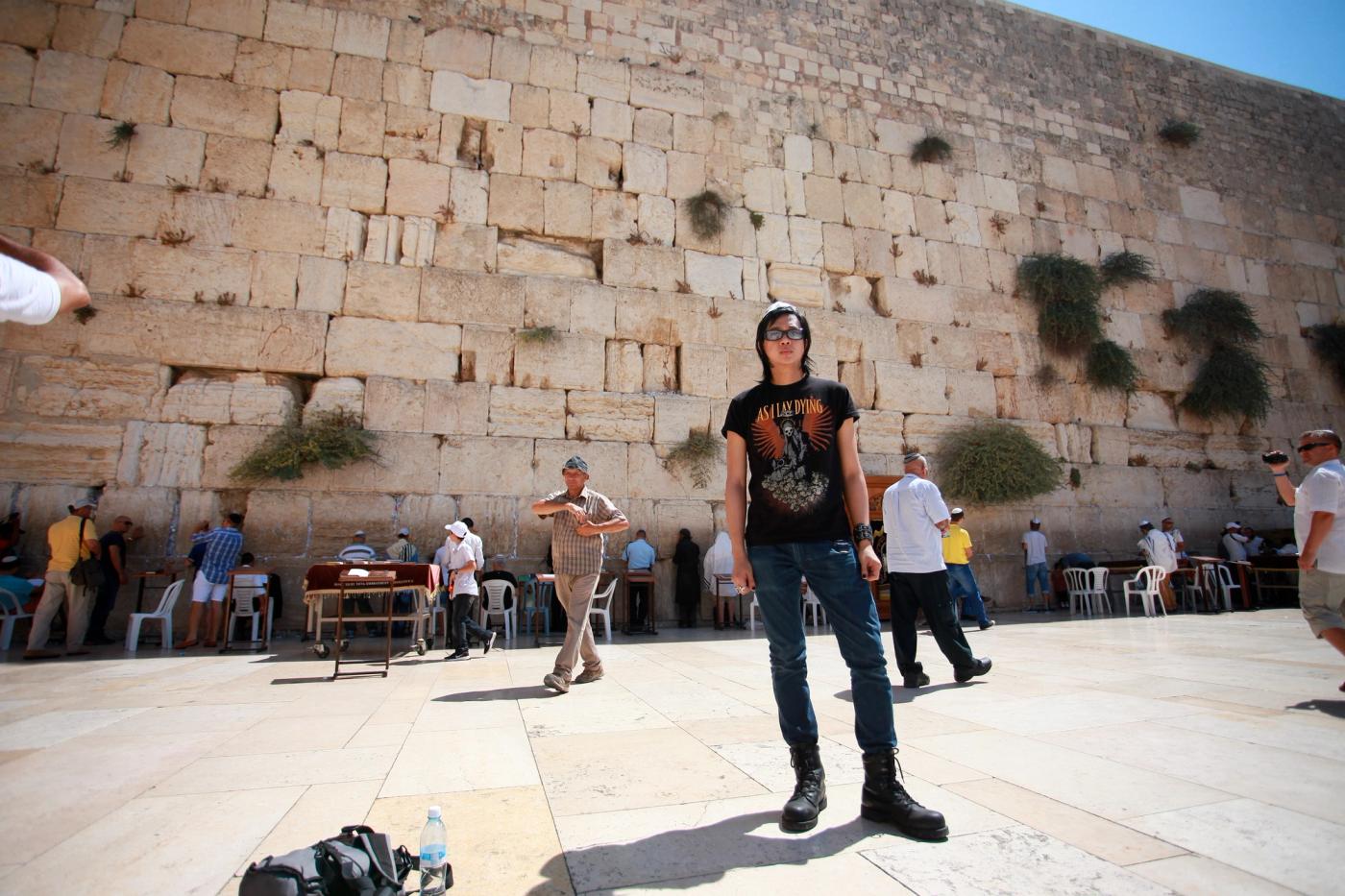 Igan D’Bayan in a black band t-shirt and boots stands in the plaza before the towering stone Western Wall in Jerusalem, worshippers praying at its base