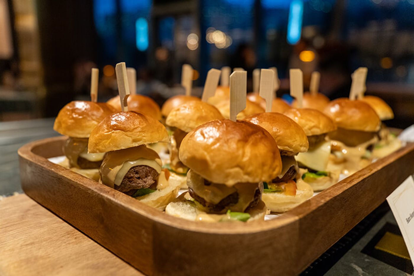 A wooden tray of H Proper cheeseburger sliders on glossy brioche buns, each speared with a pick, at Podium Social in Ortigas