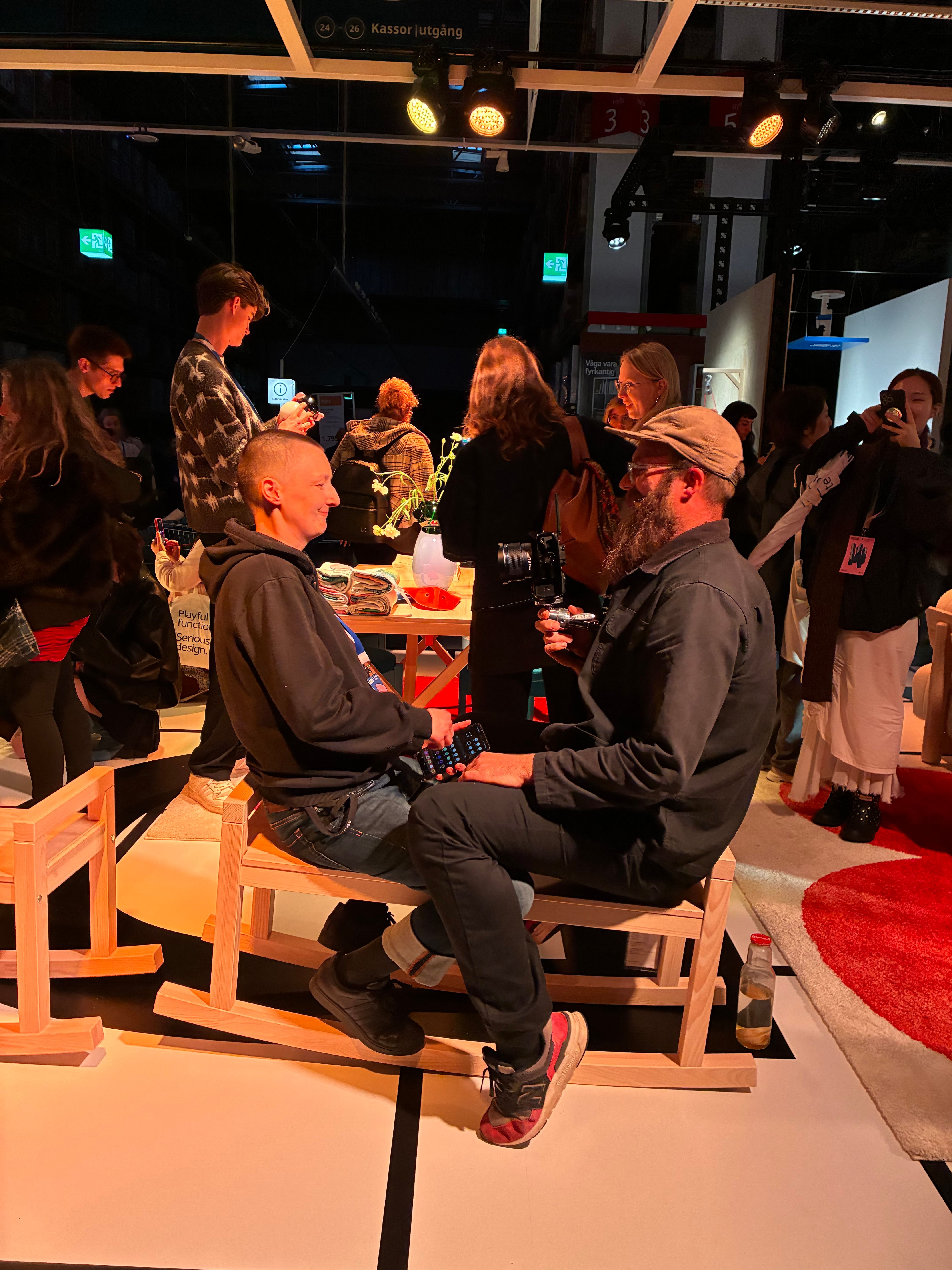 Two guests chat on small blond-wood rocking chairs inside the dim IKEA PS 2026 exhibition hall in Älmhult