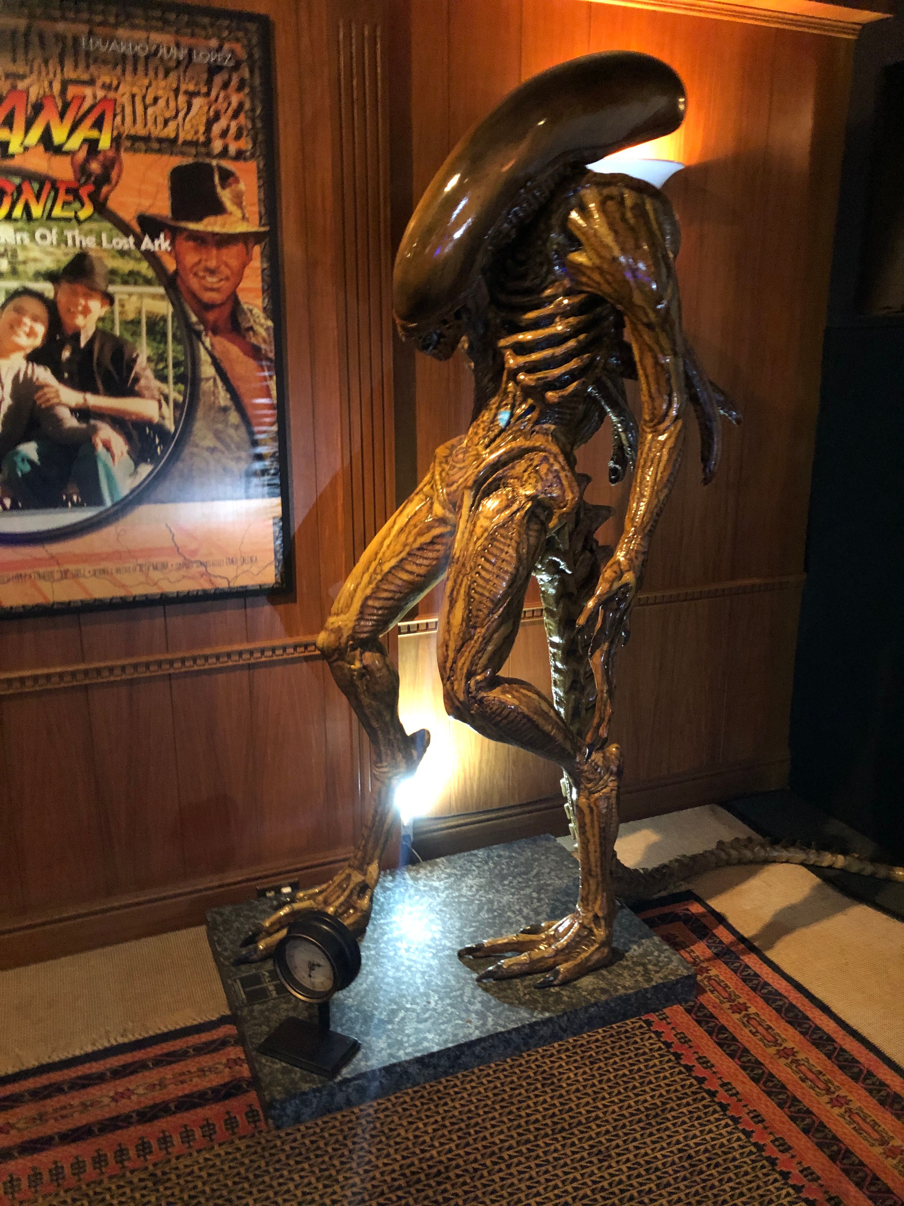 A life-size Alien xenomorph figure on a stone plinth in a wood-panelled room, beside a framed Raiders of the Lost Ark poster