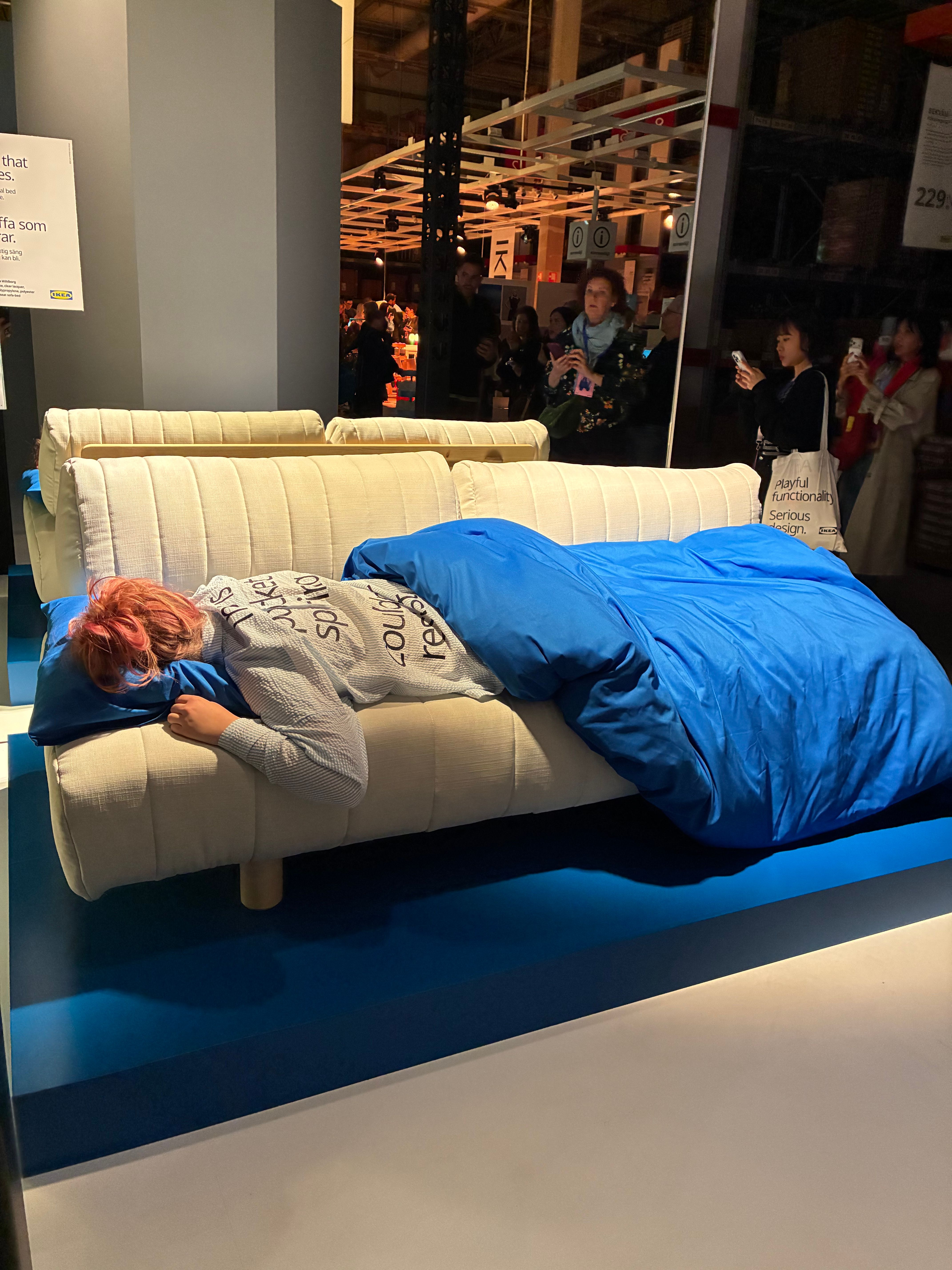 Performer sleeping under a bright blue duvet on a cream sofa bed as Democratic Design Days visitors photograph the scene