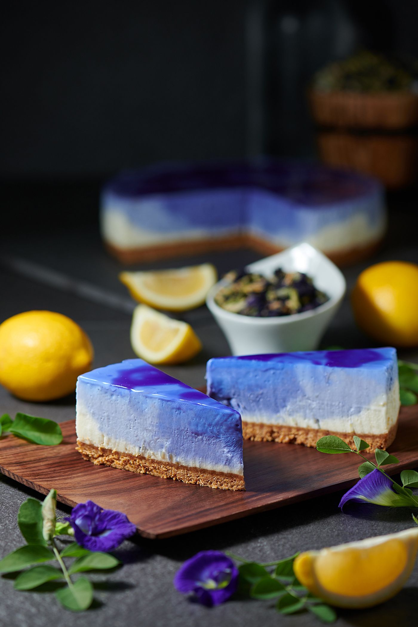 Slices of butterfly-pea lemon cheesecake with a blue-and-purple marbled top, on a wooden board with lemons and blue pea flowers