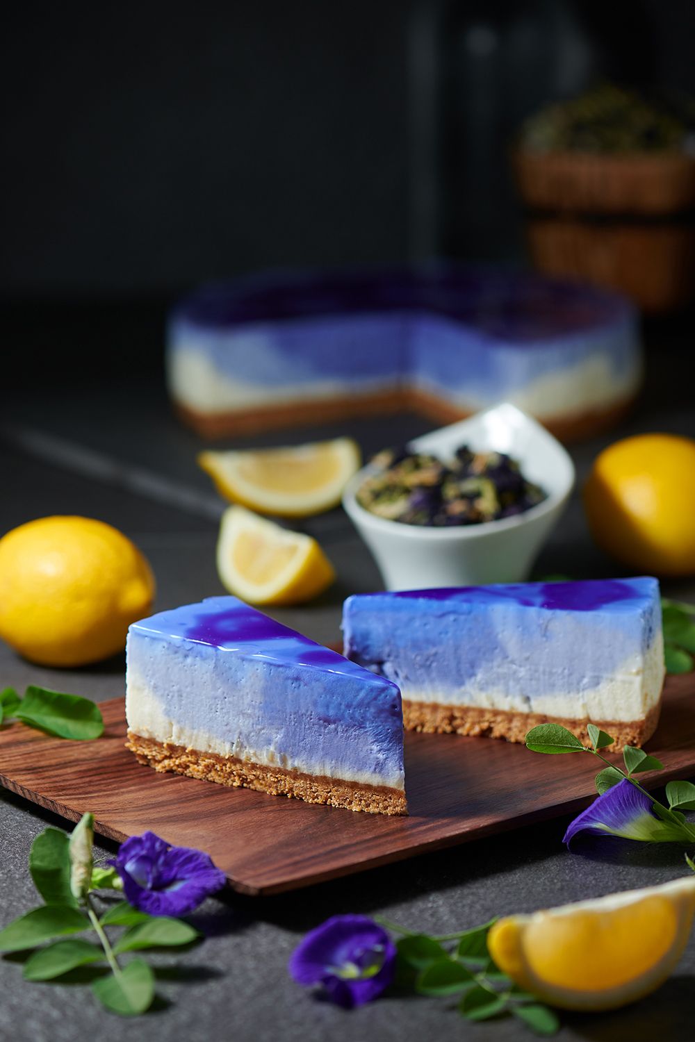 Slices of butterfly-pea lemon cheesecake with a blue-and-purple marbled top, on a wooden board with lemons and blue pea flowers