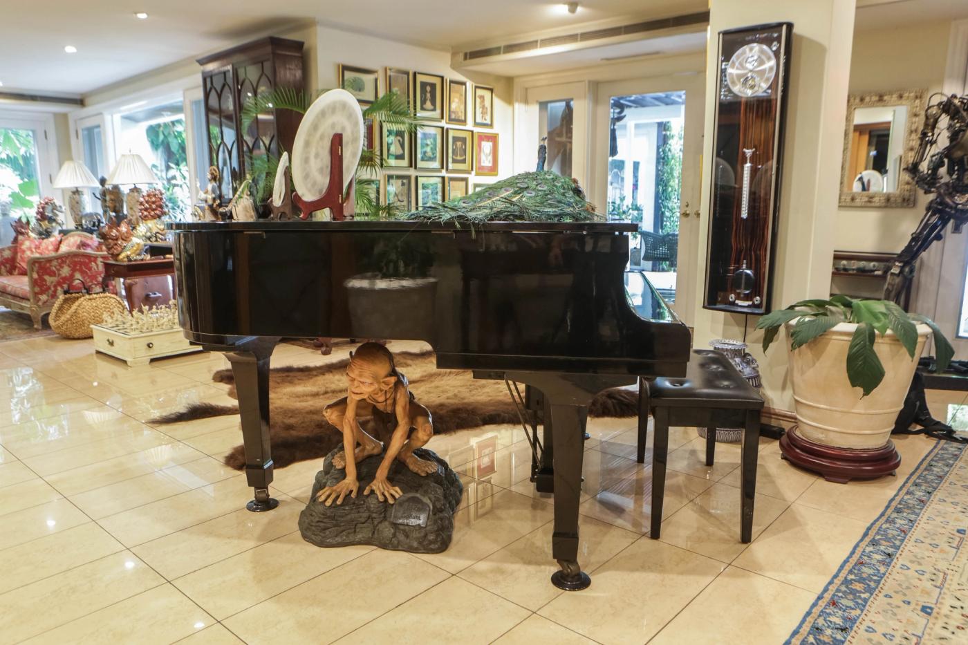 A black grand piano in a bright living room with a crouching Gollum figure beneath it and a fanned peacock on the lid