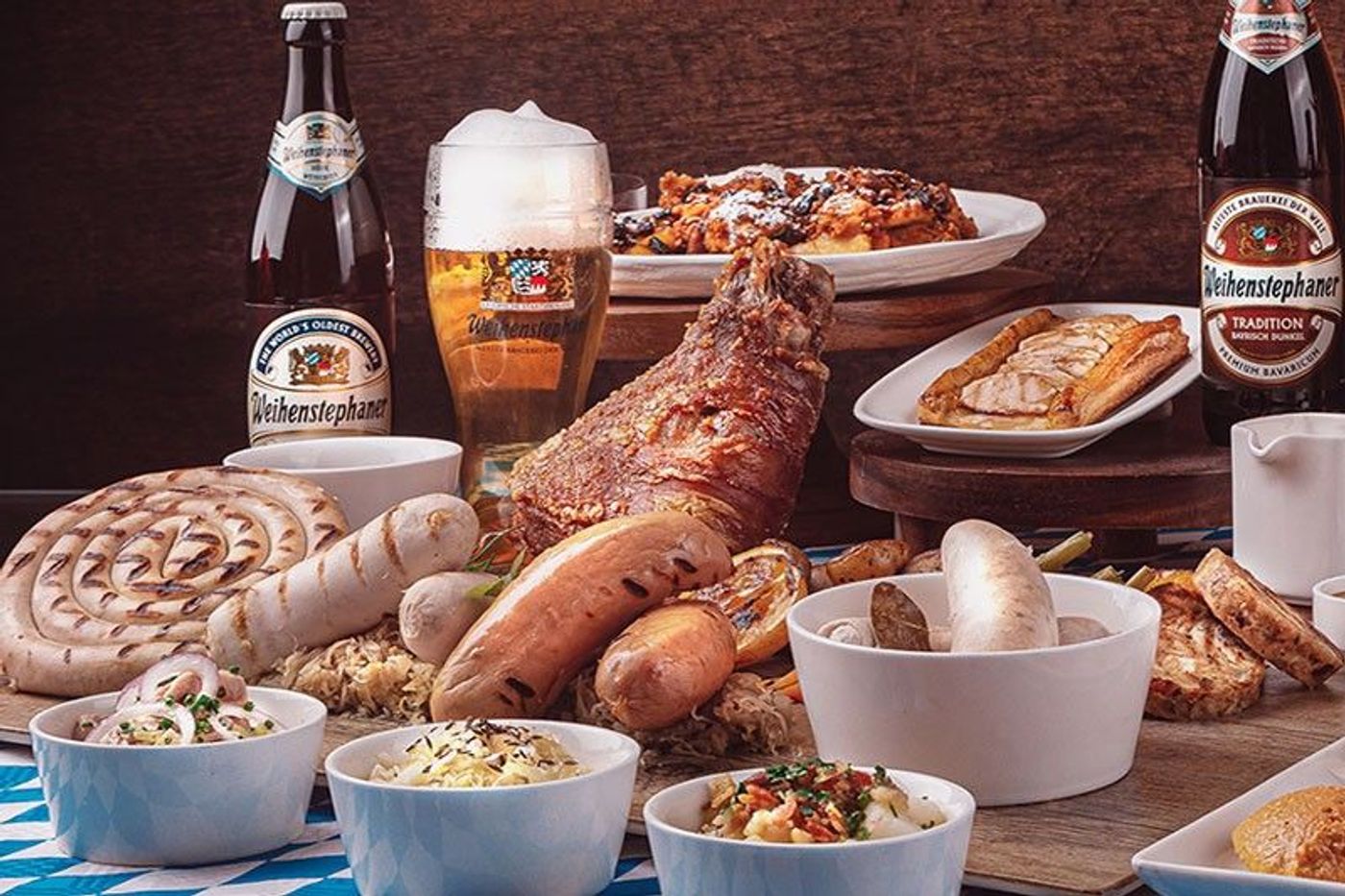 A Bavarian feast of sausages, crispy pork knuckle, sauerkraut and sides with Weihenstephan beer on a blue-checkered table