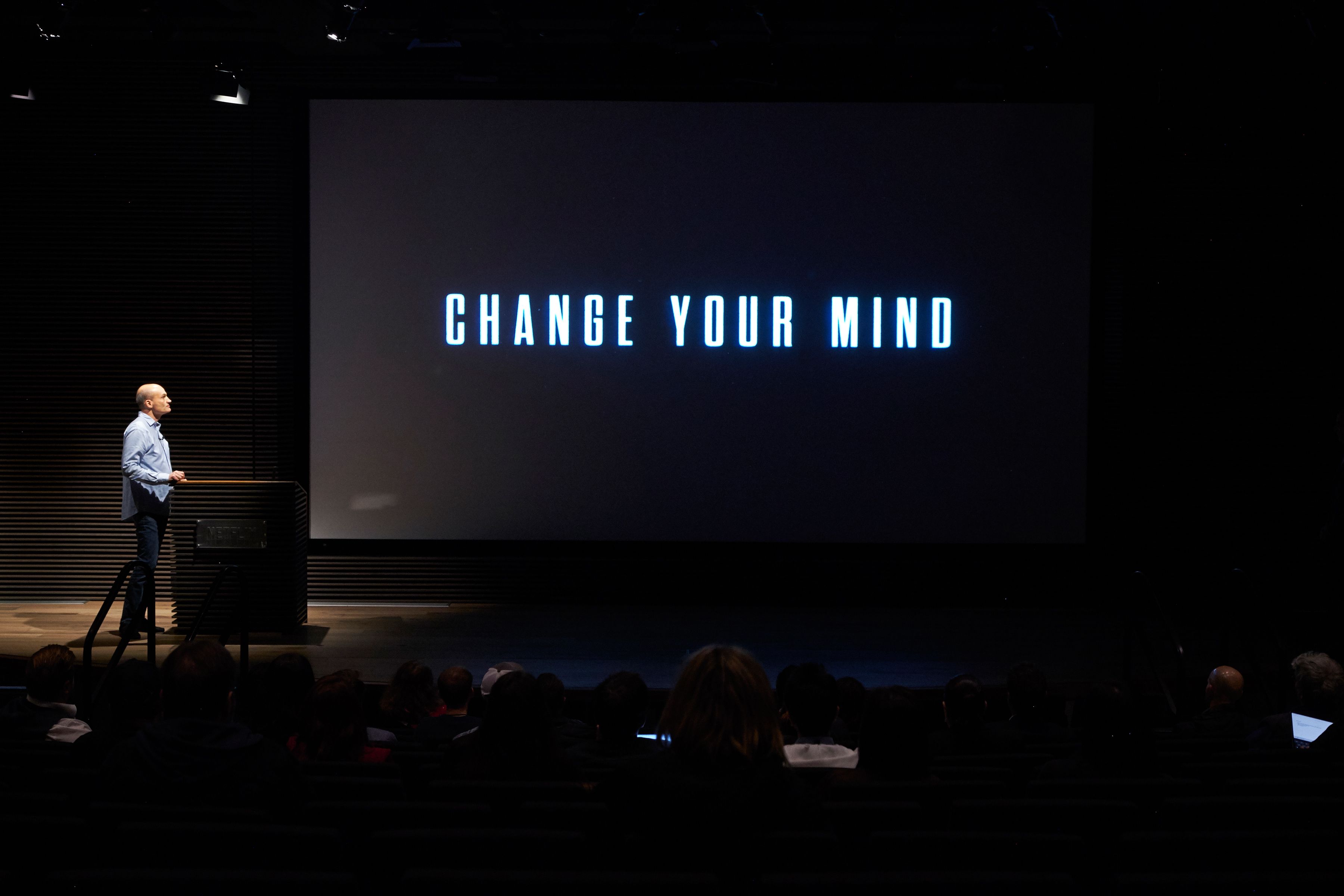 A speaker addresses a darkened auditorium as the screen reads Change Your Mind