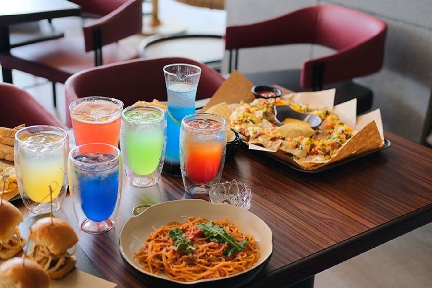 A lineup of colorful mojitos in frosted glasses on a TGI Fridays table with loaded nachos, spaghetti and mini sliders