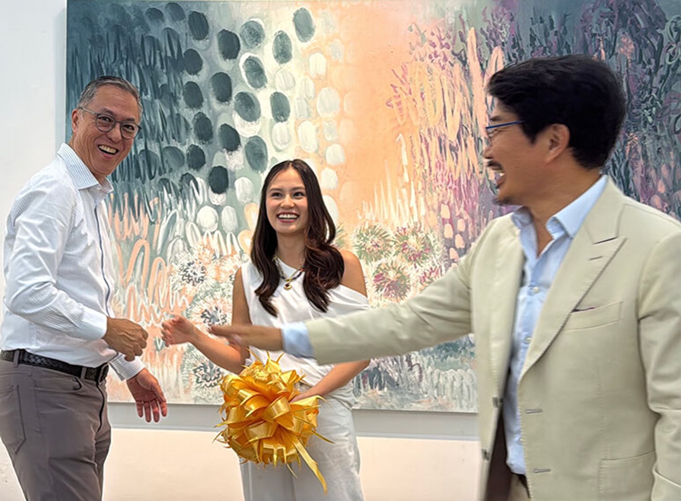 Maria Tan in a white top holding a gold ribbon bow, flanked by Michael Tan in a striped shirt and Dong Jo Chang in a beige blazer, all three laughing in front of a large botanical painting