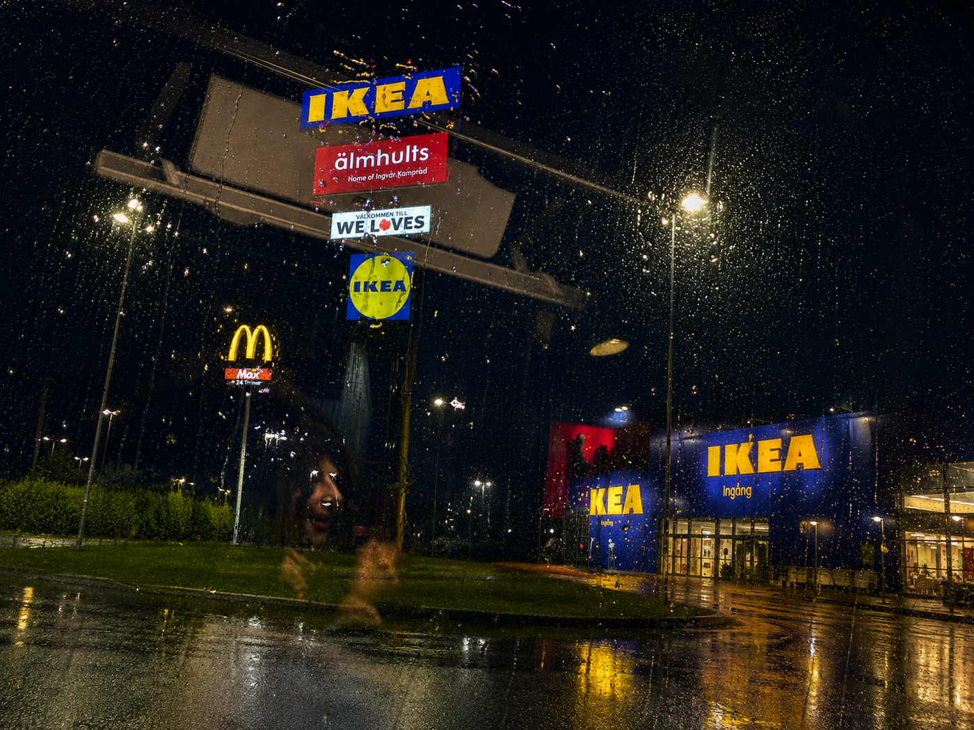 The IKEA store in Älmhult, Sweden on a rainy night, seen through a rain-beaded car window, the Home of Ingvar Kamprad sign glowing over wet pavement