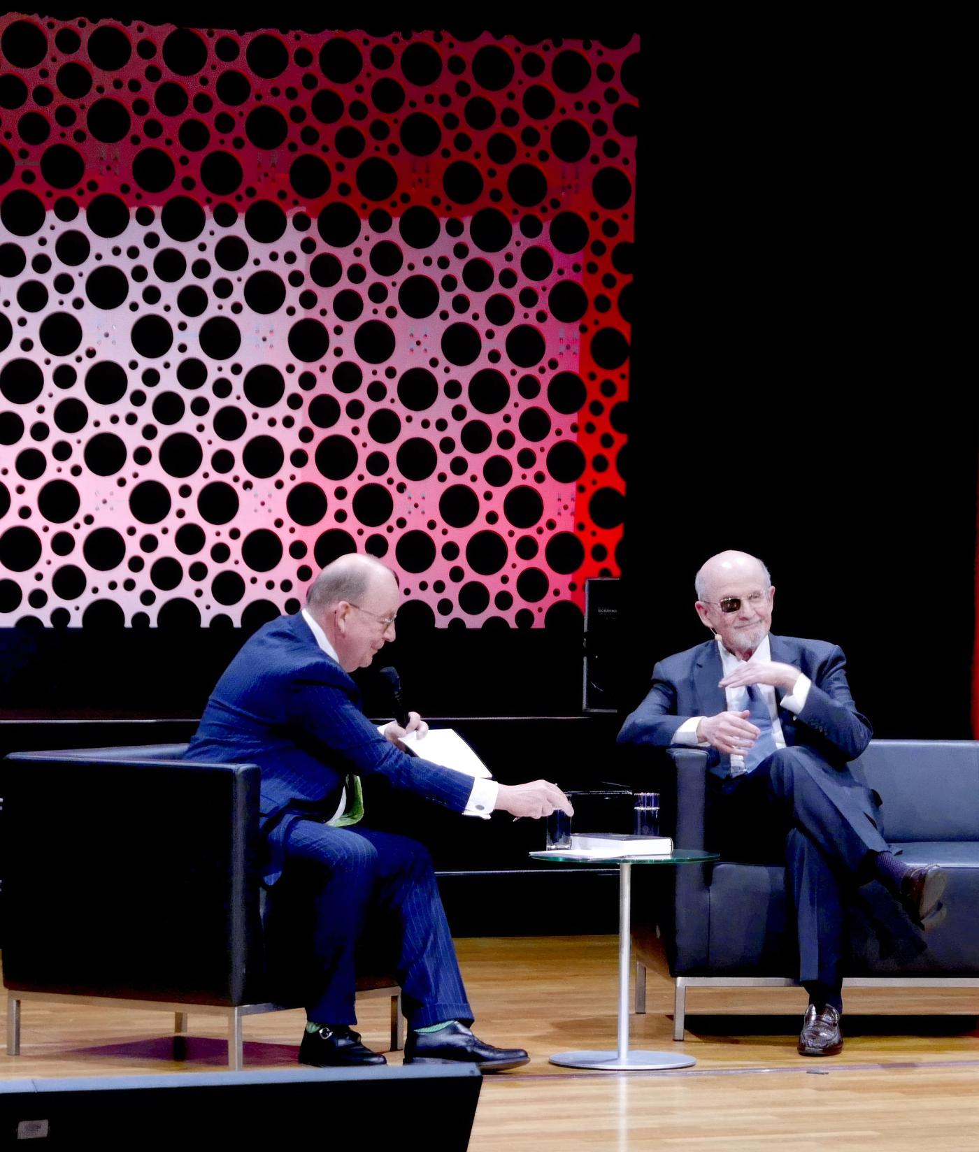 Salman Rushdie in a dark suit on a stage sofa during an onstage talk, a red dotted backdrop behind him