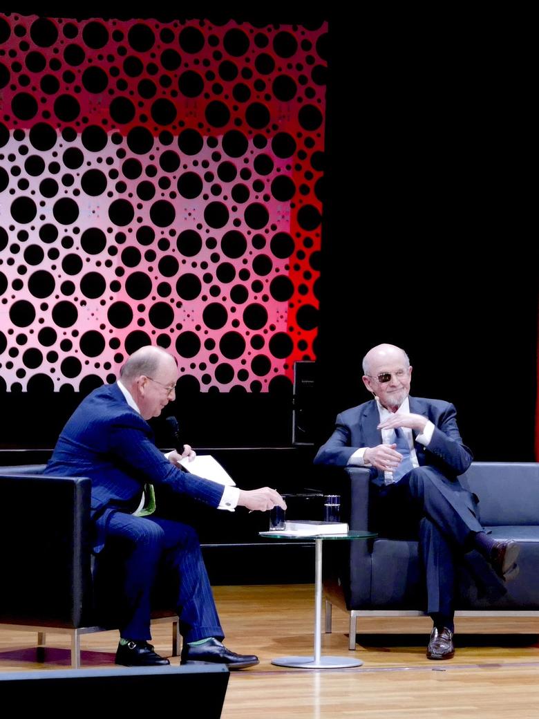 Salman Rushdie in a dark suit on a stage sofa during an onstage talk, a red dotted backdrop behind him