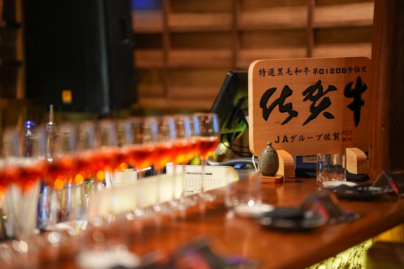 A bar counter set with rows of glasses beside a wooden Japanese signboard for Saga beef in black brush kanji