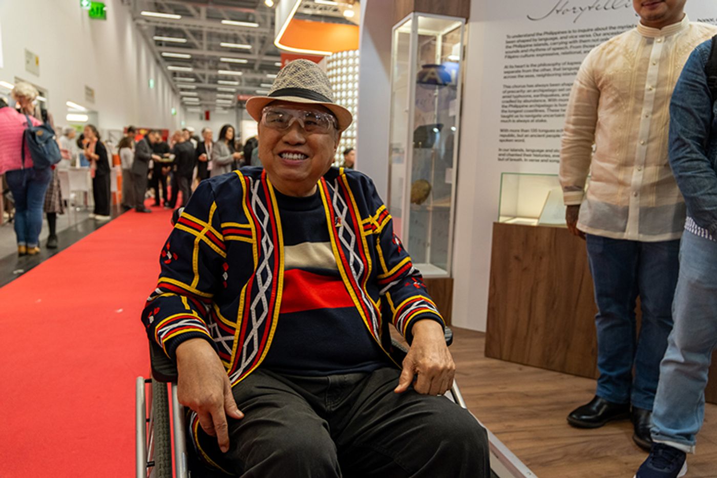 National Artist Virgilio Almario smiling in a colorful woven jacket and hat, seated in a wheelchair at the fair