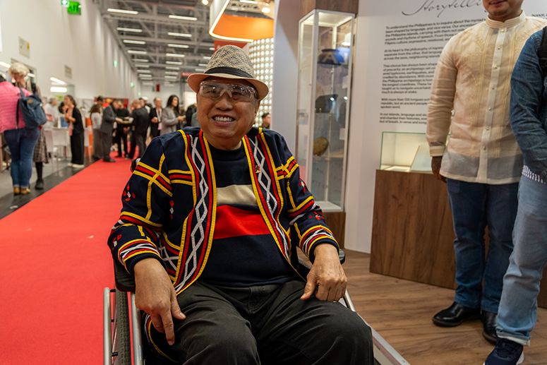 National Artist Virgilio Almario smiling in a colorful woven jacket and hat, seated in a wheelchair at the fair