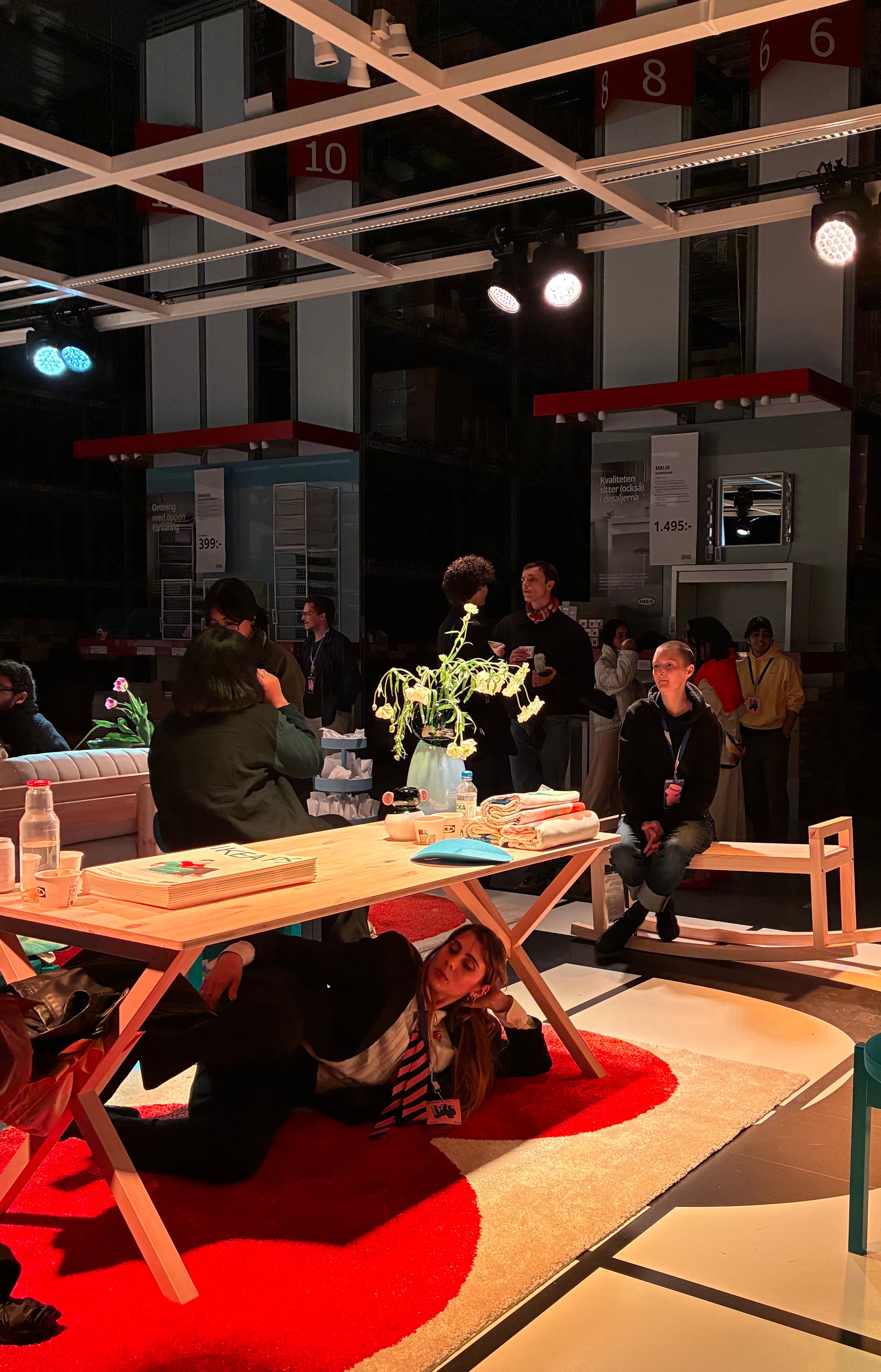 Performer reclining under a pine trestle table on a red and white rug during a Democratic Design Days showcase inside the Älmhult warehouse