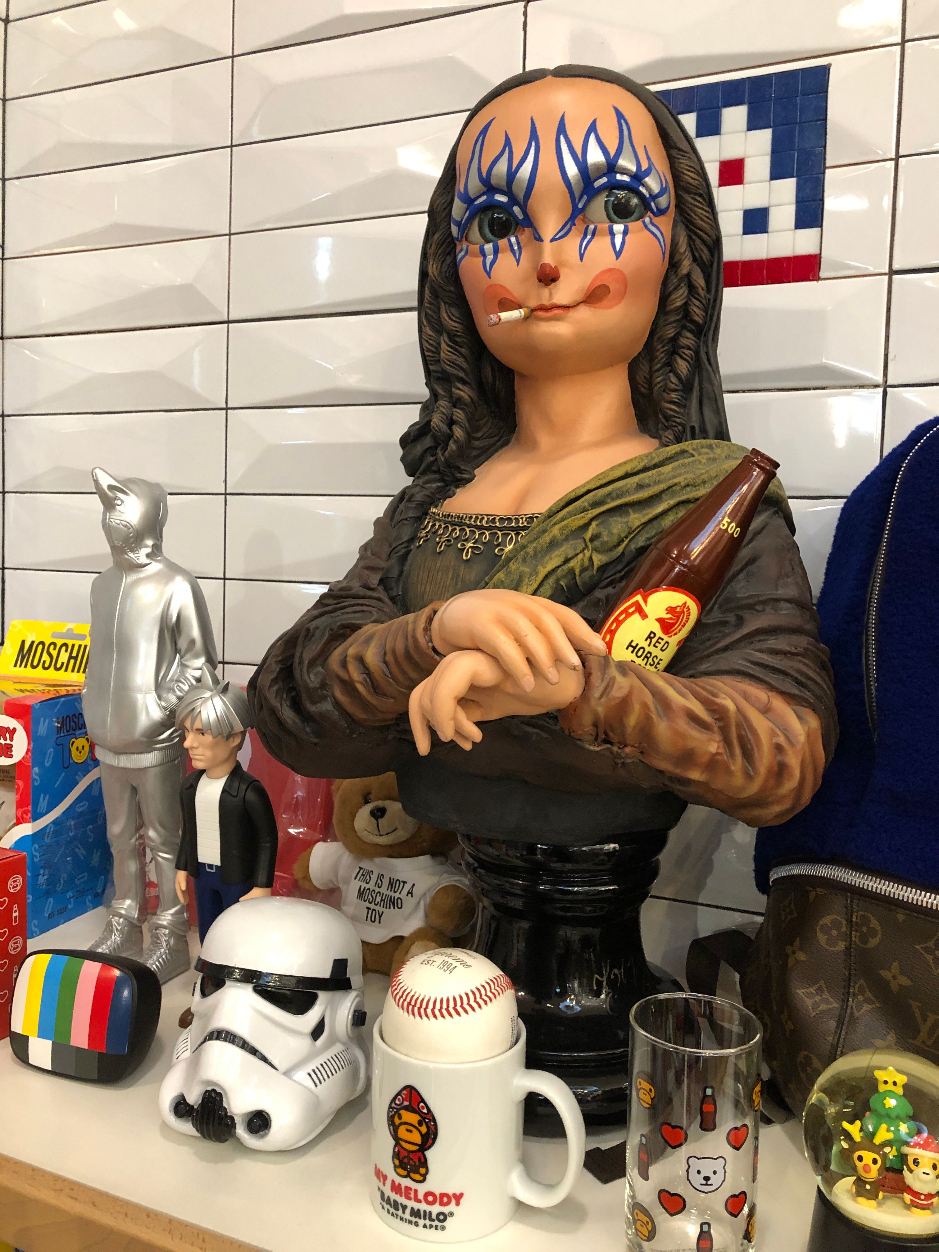 A Mona Lisa bust with blue face paint, a cigarette in her mouth and a Red Horse beer bottle, among a Stormtrooper helmet, Moschino boxes and a Baby Milo mug