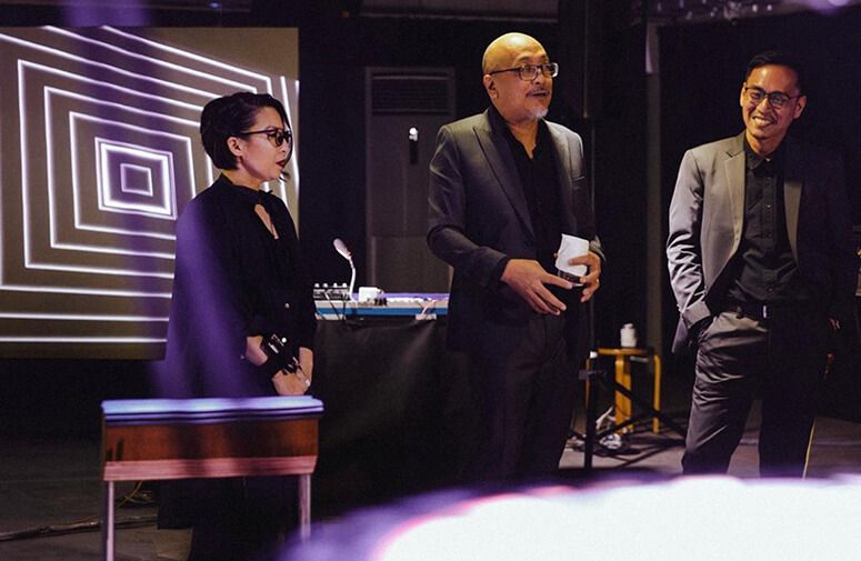 Christina Dy, Tad Ermitaño, and Marco Ortiga of Somatosonic standing in a dark room under stage lights, speaking in front of a concentric square projection