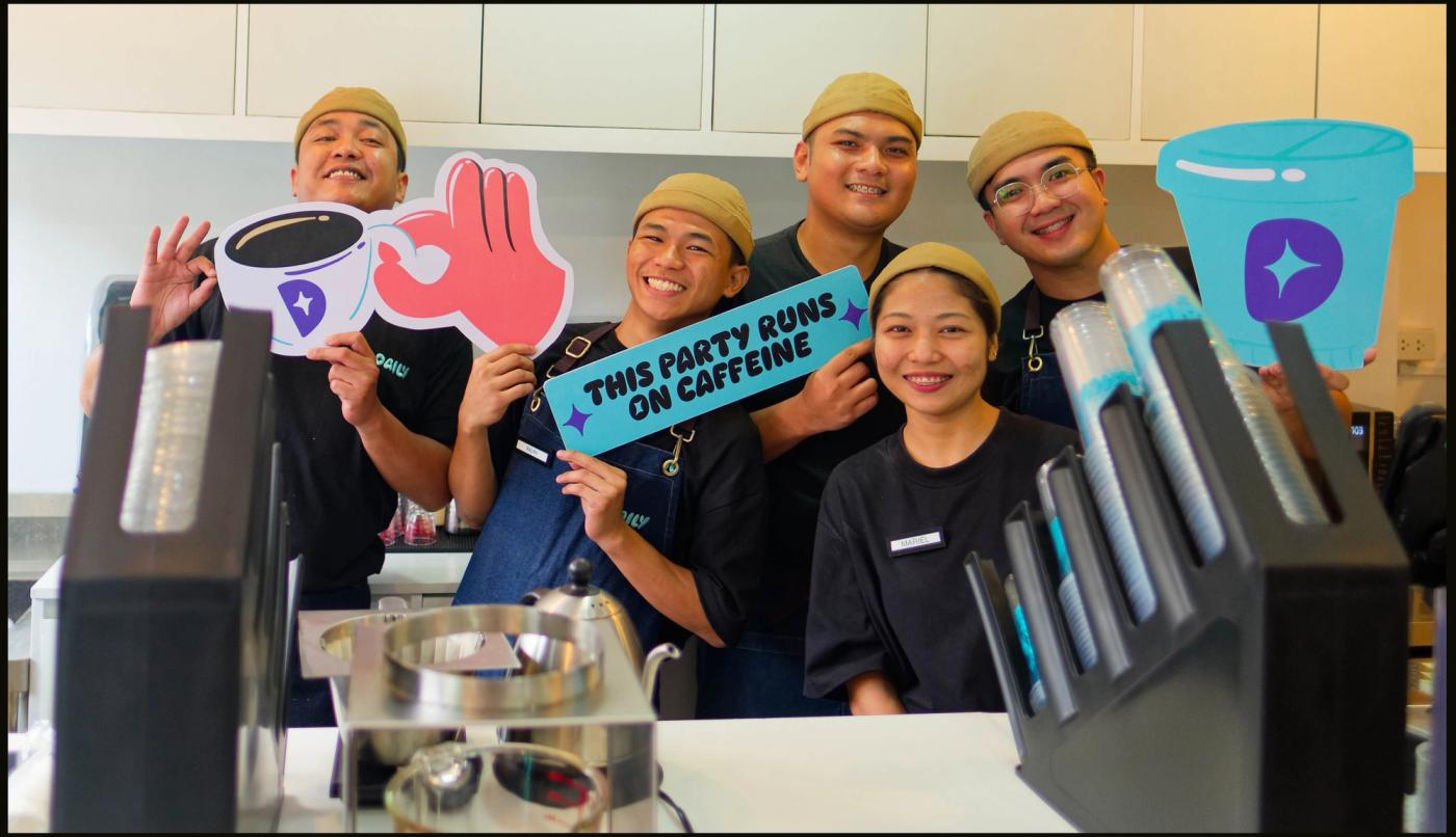 Five Daily by Bo’s Coffee staff in tan caps grin behind the counter holding coffee-cup cutouts and a ‘This party runs on caffeine’ sign
