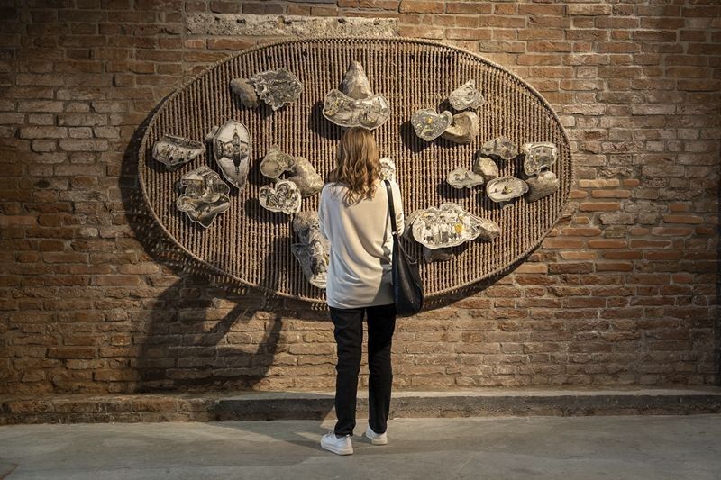 A visitor studies Ronald Ventura’s oval woven wall piece studded with stone-like relief fragments on a brick wall in Venice