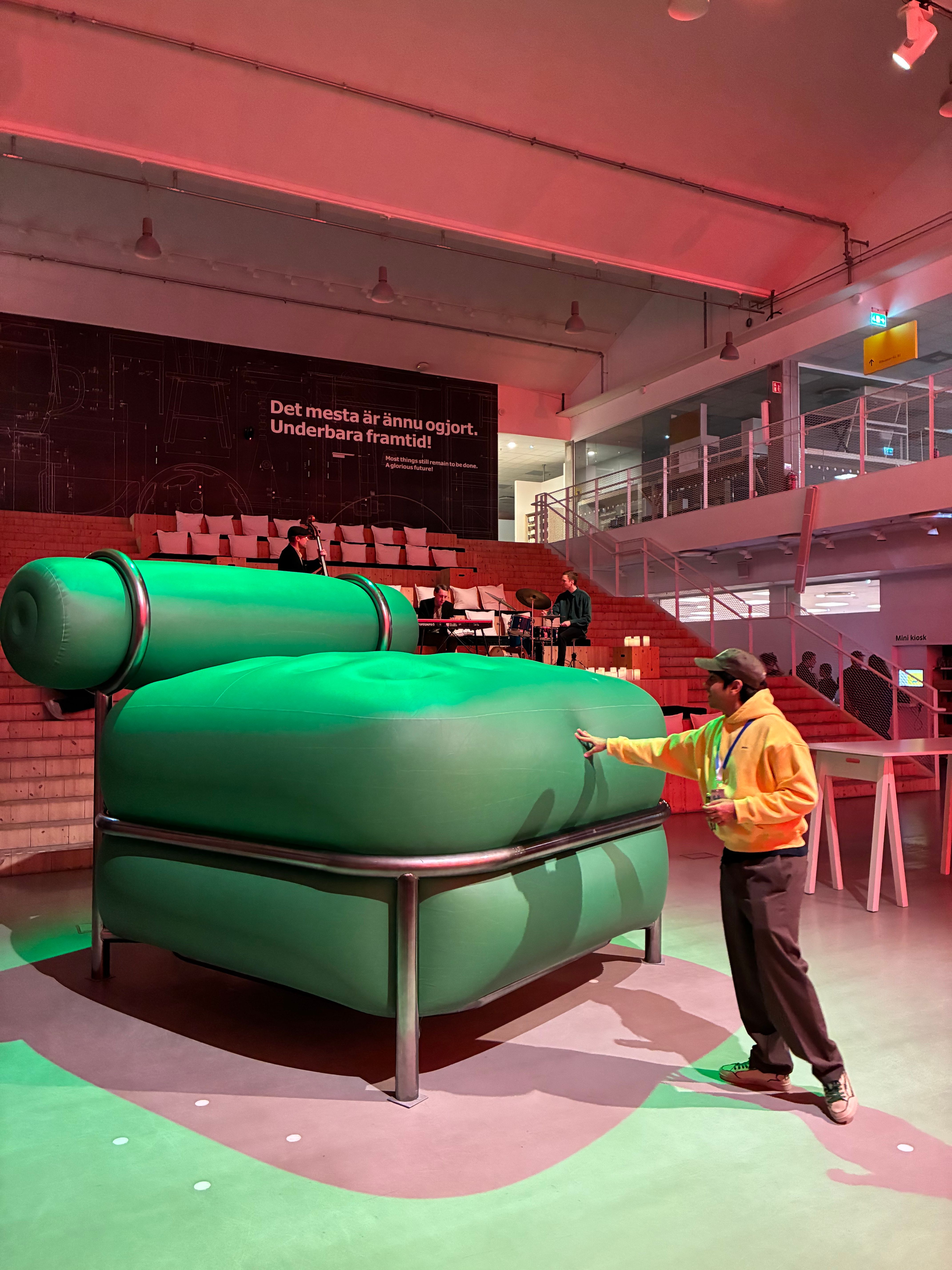 Visitor touching an oversized green inflatable armchair on a steel frame while a band plays on stepped seating behind