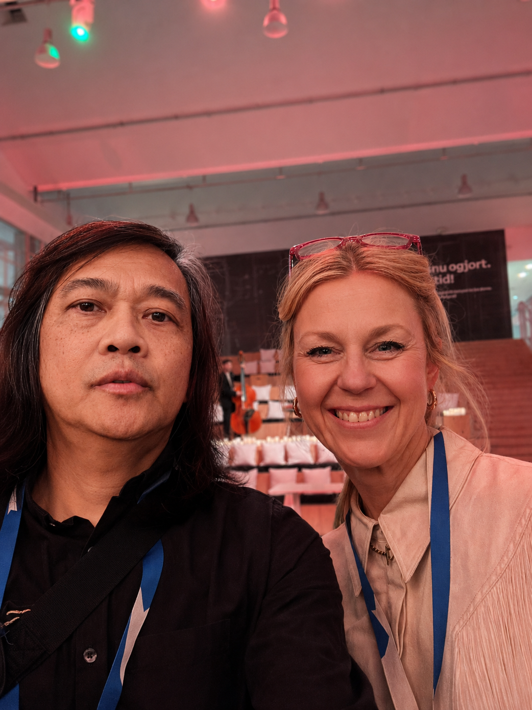 Two attendees take a selfie in a pink-lit event hall with tiered cushion seating and a double bass on stage behind them