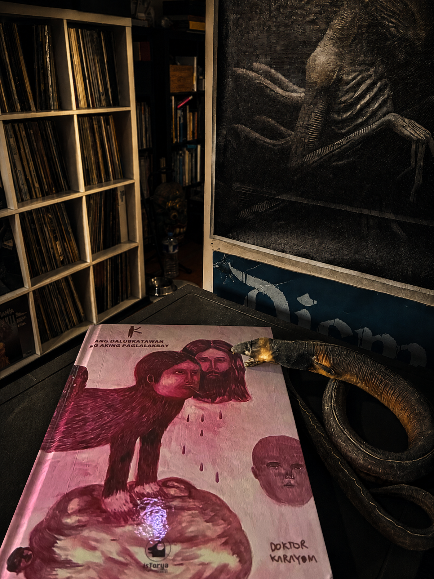 Doktor Karayom’s book Ang Dalubkatawan ng Aking Paglalakbay, its crimson two-headed figure on the cover, beside a carved serpent and crates of vinyl records