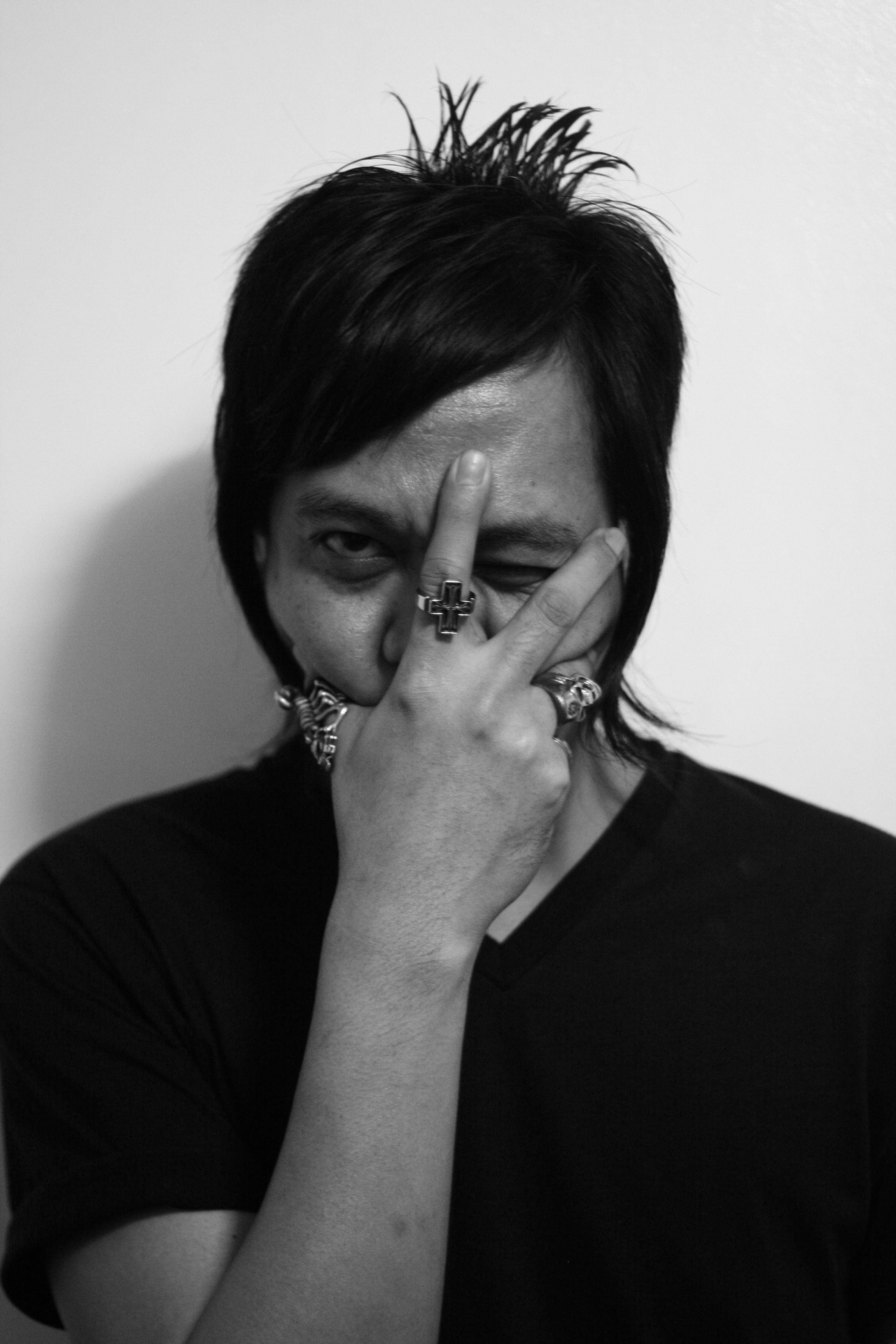 Igan D'Bayan in a black and white portrait, wearing a black V-neck T-shirt with dark layered hair falling across one eye, one hand raised to his face with two fingers parted over his eyes and a cross-shaped silver ring on one of them, staring back through the gap