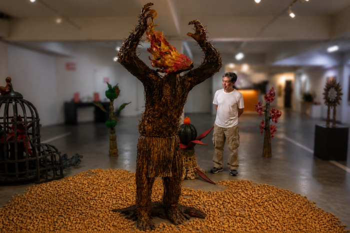 Bob Feleo in a white shirt looking up at his life-size bark-textured figure with a flame for a head, arms raised over a wide circle of small clay balls on the gallery floor