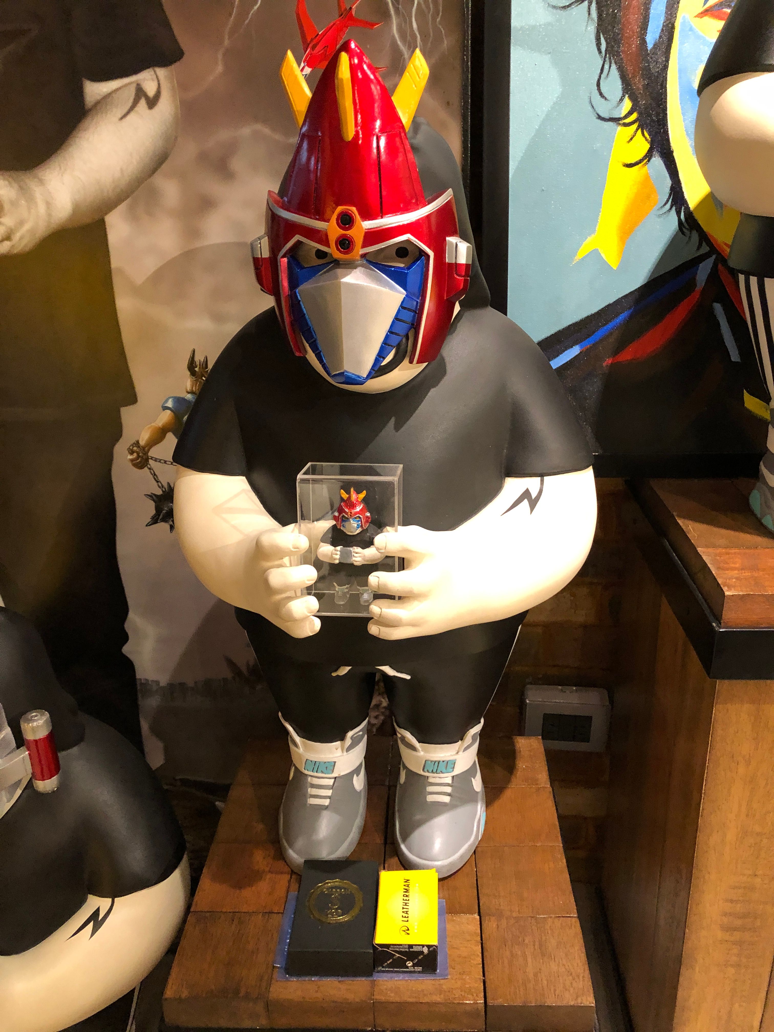 An art toy of a heavy-set figure in a Voltes V helmet and black shirt, cradling a boxed miniature of the same robot and wearing grey Nike Mag sneakers