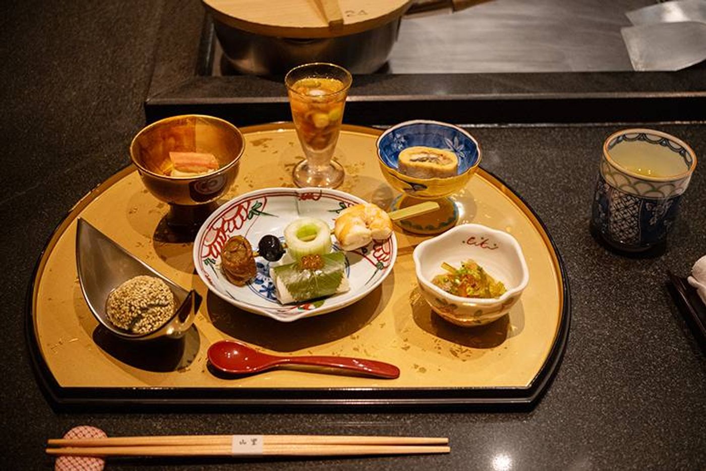 Kaiseki appetizers on a gold lacquered tray: small ceramic dishes, a sesame-dusted bite and a chilled glass