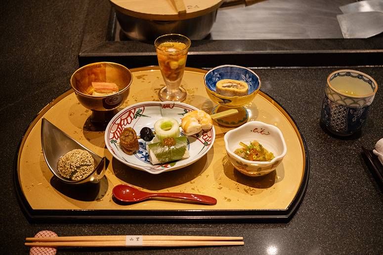 Kaiseki appetizers on a gold lacquered tray: small ceramic dishes, a sesame-dusted bite and a chilled glass