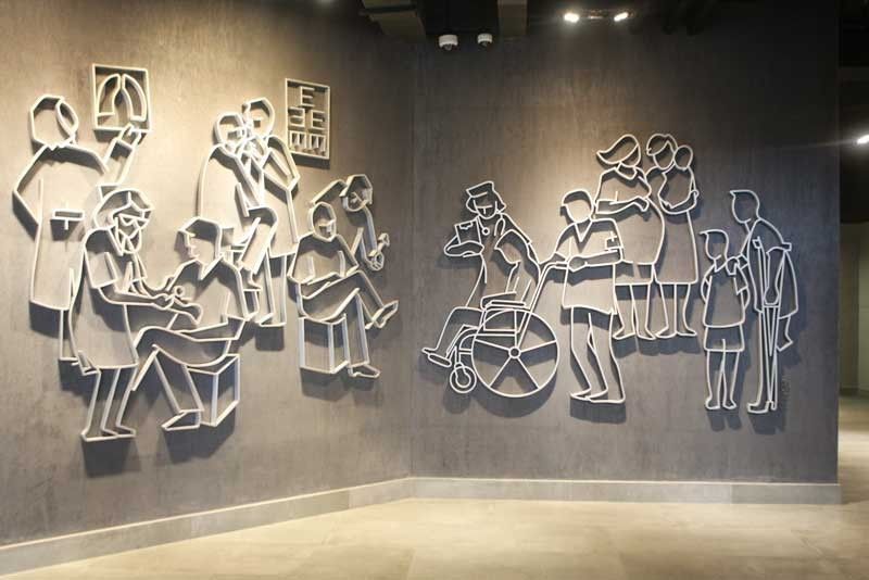 White outline reliefs of clinic scenes across two grey walls: a doctor with a patient, an eye chart, a figure in a wheelchair and a child on crutches