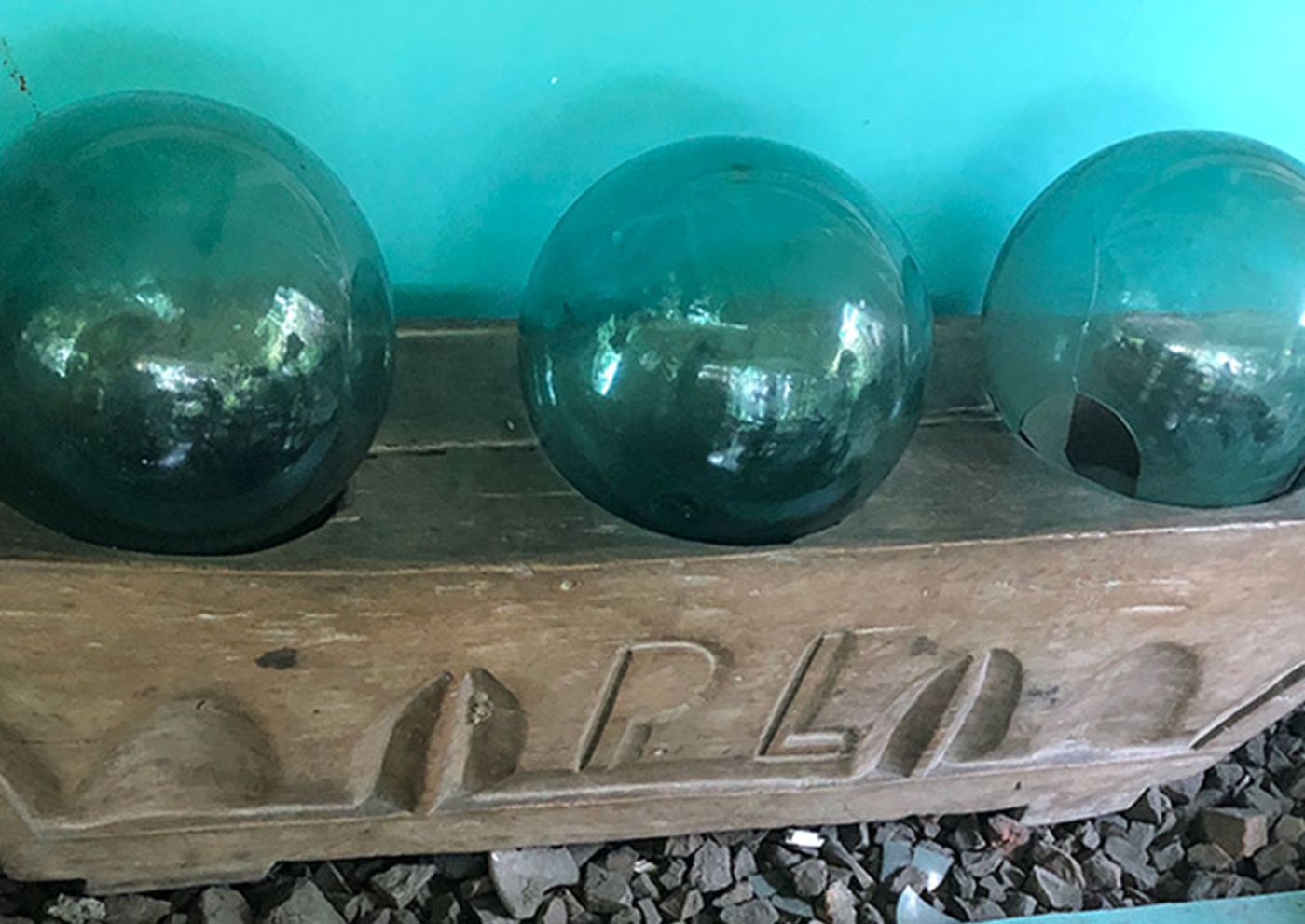 Three green glass fishing floats resting on a salvaged carved wooden beam, the wall behind them painted turquoise