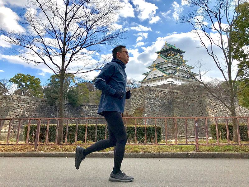 Emerson Yao running in a navy jacket and tights on a path below Osaka Castle, bare trees and a stone rampart behind him