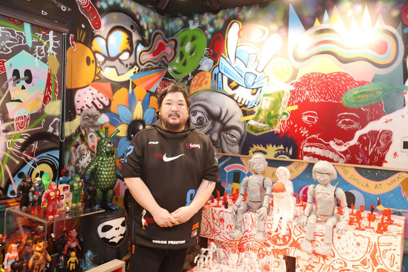 Bigboy Cheng in a black Nike shirt in front of a wall mural of cartoon faces, beside shelves of vinyl figures and white robot sculptures
