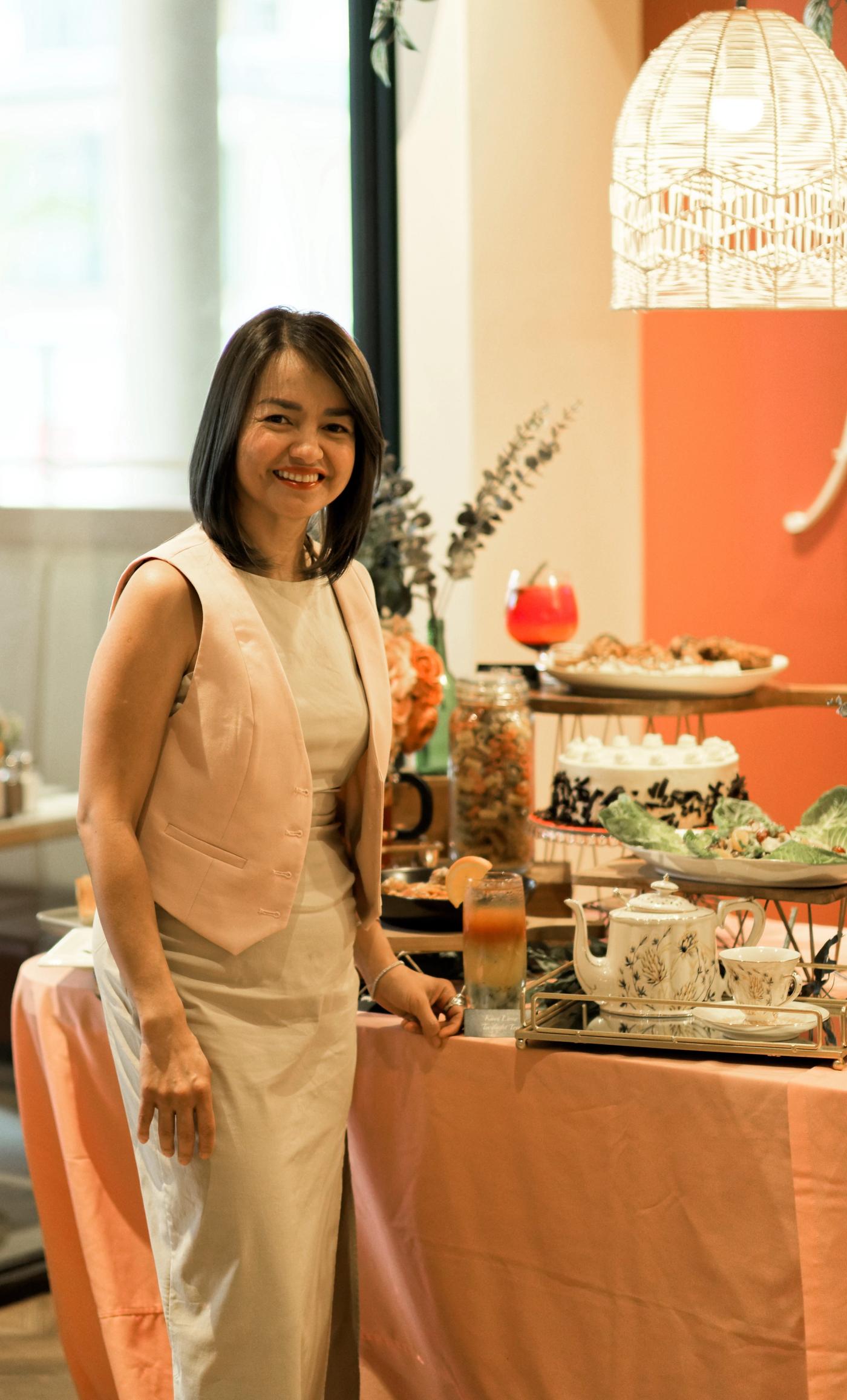 RC Tiongson in a sleeveless beige dress smiles beside a Secret Recipe dessert display of cakes and a pink drink