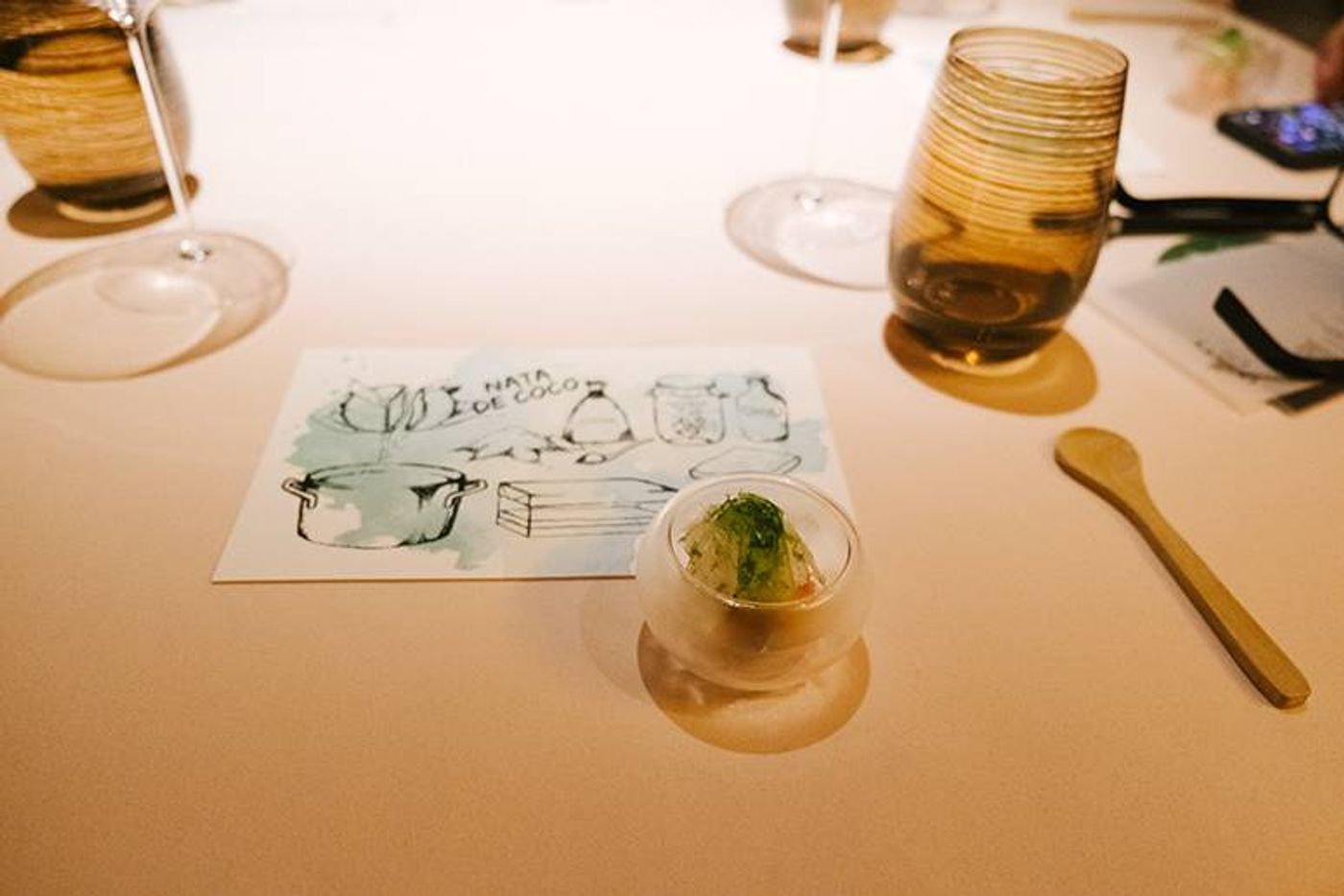 A glass sphere of buko salad sits on an illustrated nata de coco card beside a wooden spoon and wine glasses at Chef Chele Gonzalez’s coconut dinner