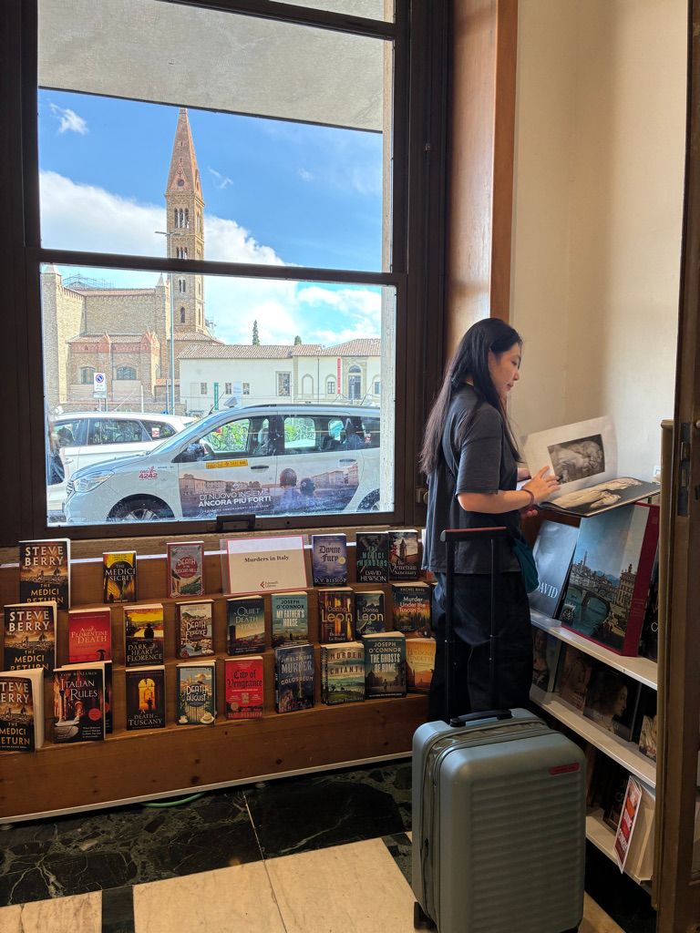 Avee Navarro Tan reading in a Florence bookshop by a window framing a brick bell tower, a silver cabin suitcase beside her and a shelf of Murders in Italy crime novels below