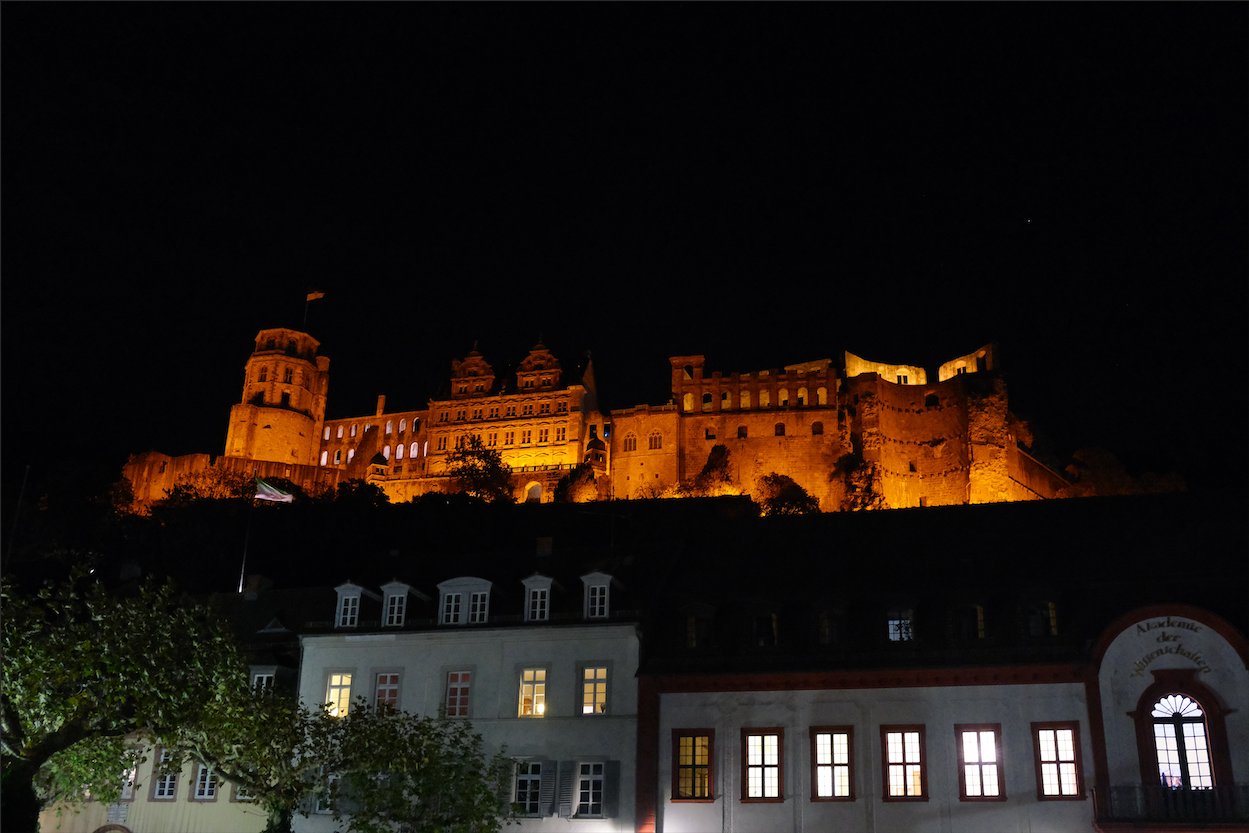 Heidelberg Castle floodlit amber against the night sky, its towers and broken walls rising above the pale facades and dark rooftops of the old town below