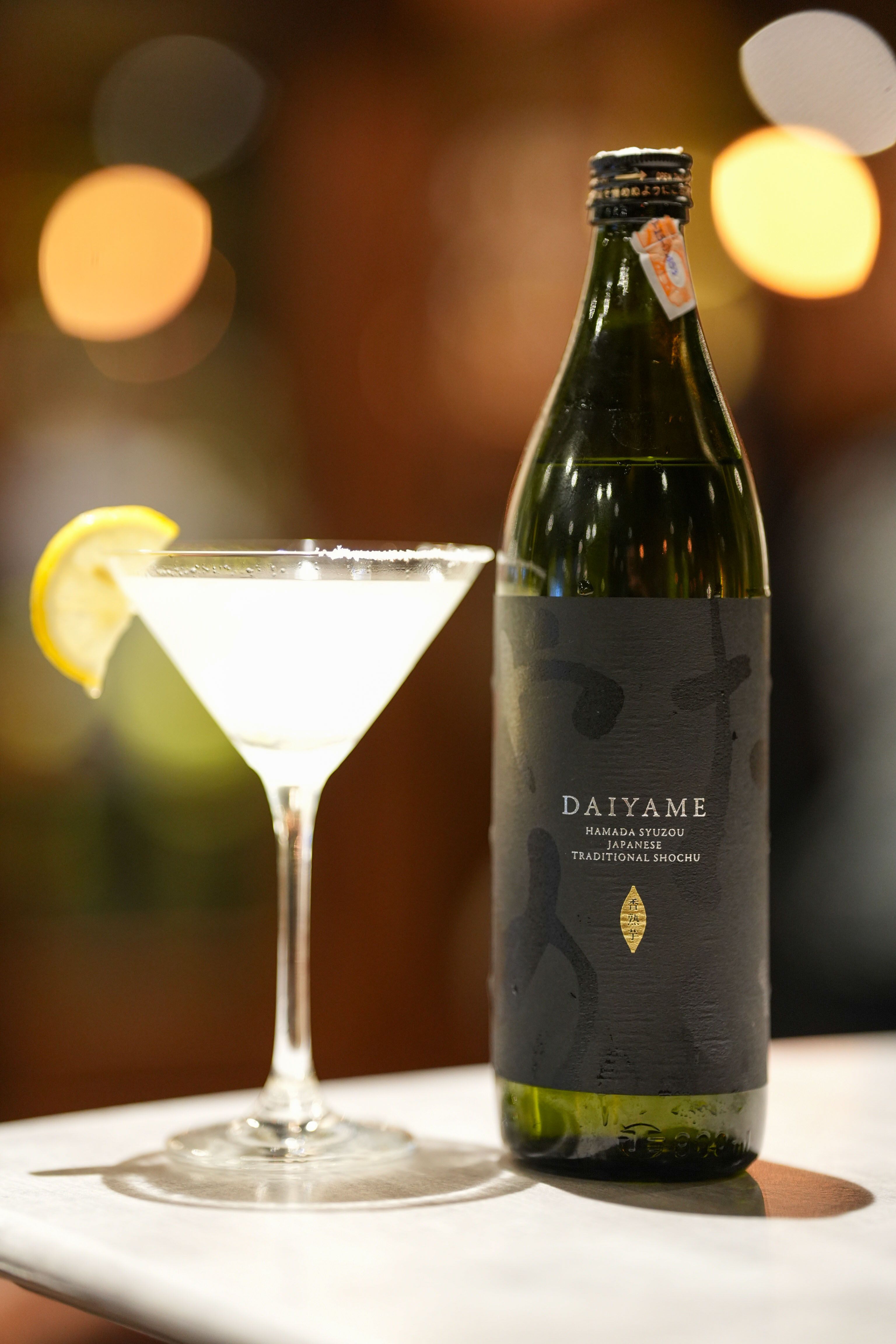 A pale cocktail garnished with lemon beside a bottle of Daiyame Japanese shochu on a marble table