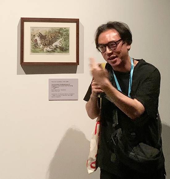 Artist Ho Tzu Nyen in glasses gesturing beside a small framed print on a white gallery wall