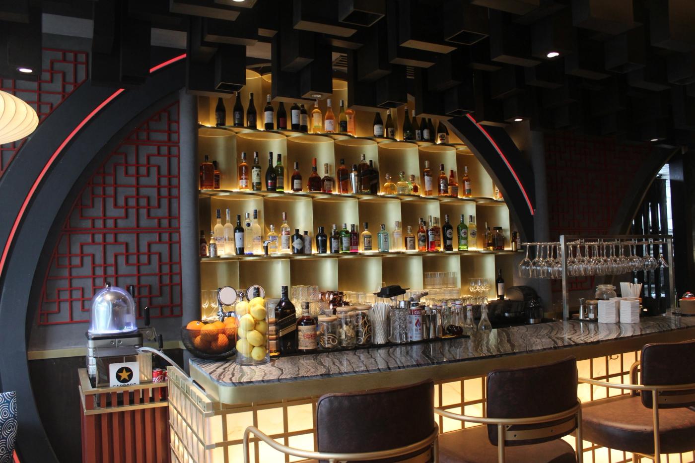 A contemporary bar counter inside Red Lotus restaurant featuring traditional Chinese red lattice patterns, a golden back bar shelf stocked with bottles, and dark wooden block ceiling structures