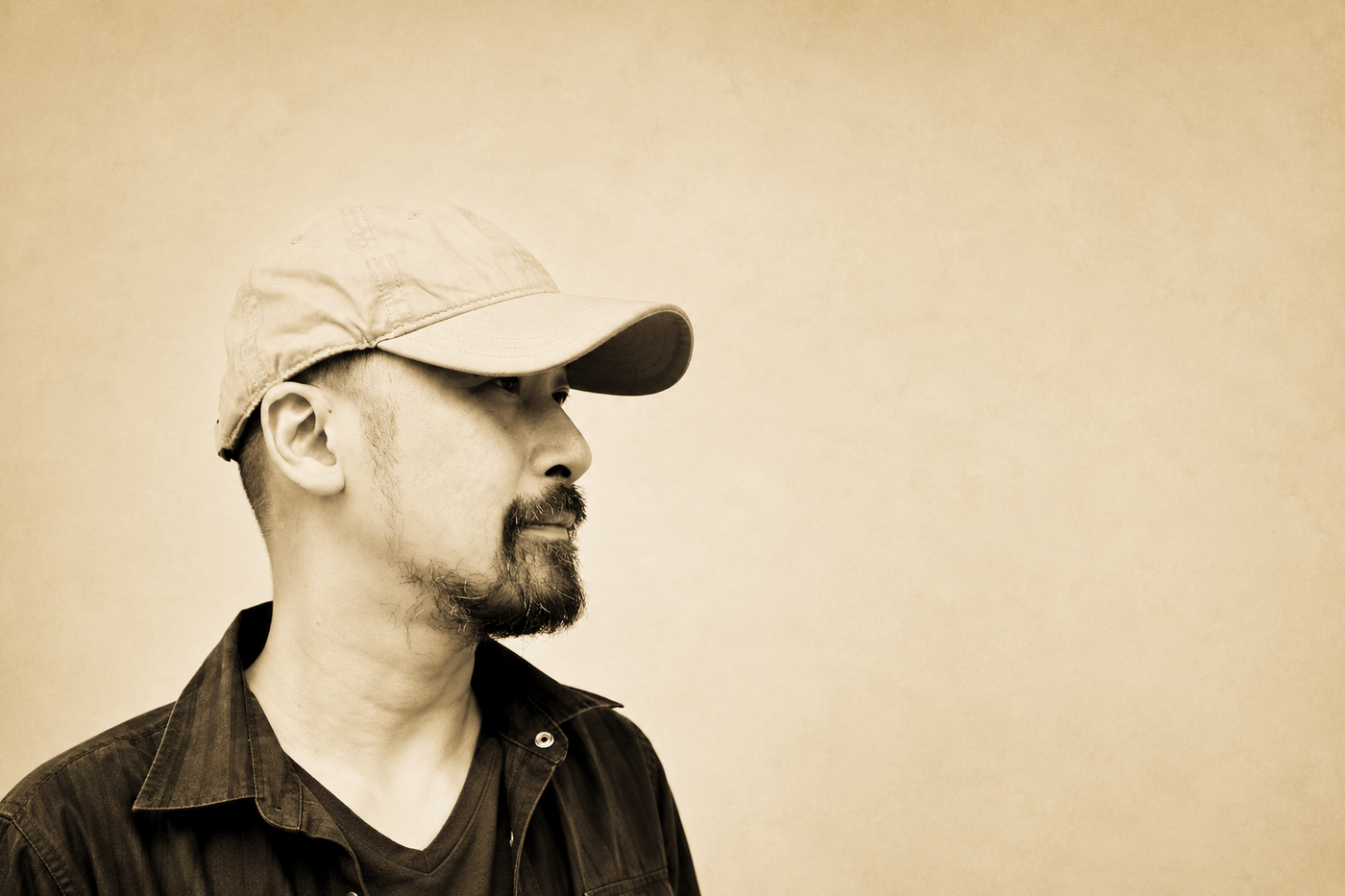 Artist Manuel Ocampo in profile, bearded and in a pale baseball cap and dark work shirt, against a plain sepia-toned wall