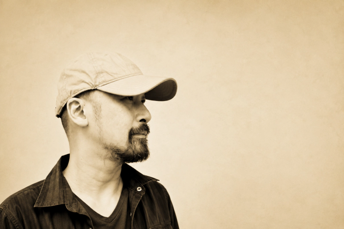 Artist Manuel Ocampo in profile, bearded and in a pale baseball cap and dark work shirt, against a plain sepia-toned wall