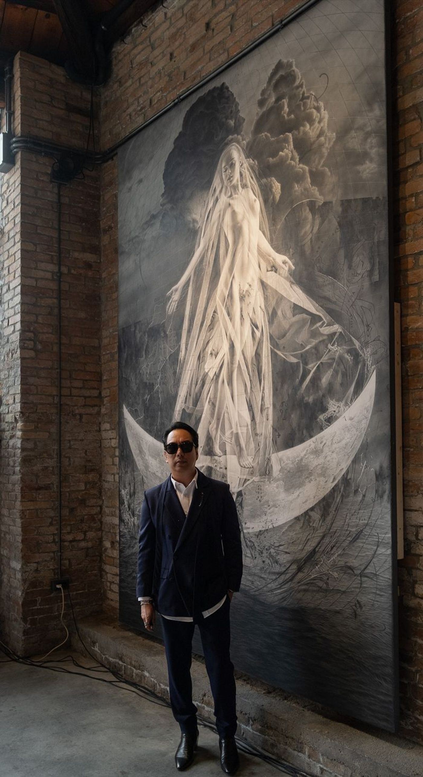 Ronald Ventura in a navy suit and sunglasses stands before his towering monochrome “LUNA” work of a veiled figure on a crescent moon