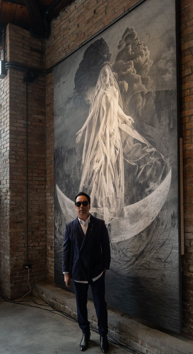 Ronald Ventura in a navy suit and sunglasses stands before his towering monochrome “LUNA” work of a veiled figure on a crescent moon