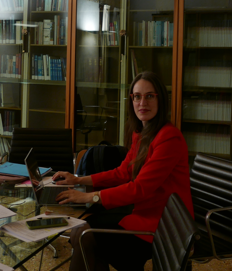 A woman in glasses and a red top works on a laptop at a desk in a library lined with glass-front book cabinets