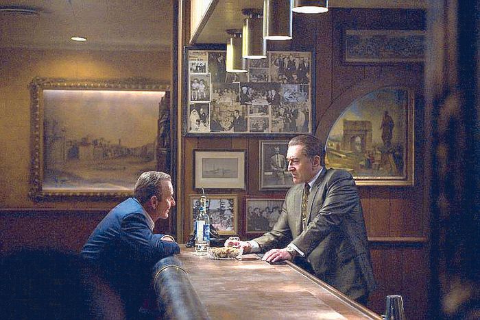 Joe Pesci and Robert De Niro talk across a dimly lit wooden bar lined with framed photographs